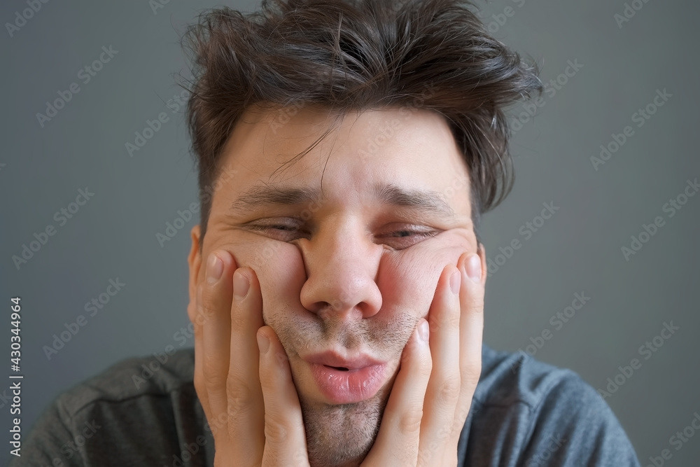 Tired boring shaggy young man is looking at camera on grey background. He is keeping his head by hands. Depression, apathy, and mental illness in the modern world. Middle age crisis. Tired boring shaggy young man is looking at camera on grey background. He is keeping his head by hands. Depression, apathy, and mental illness in the modern world. Middle age crisis.