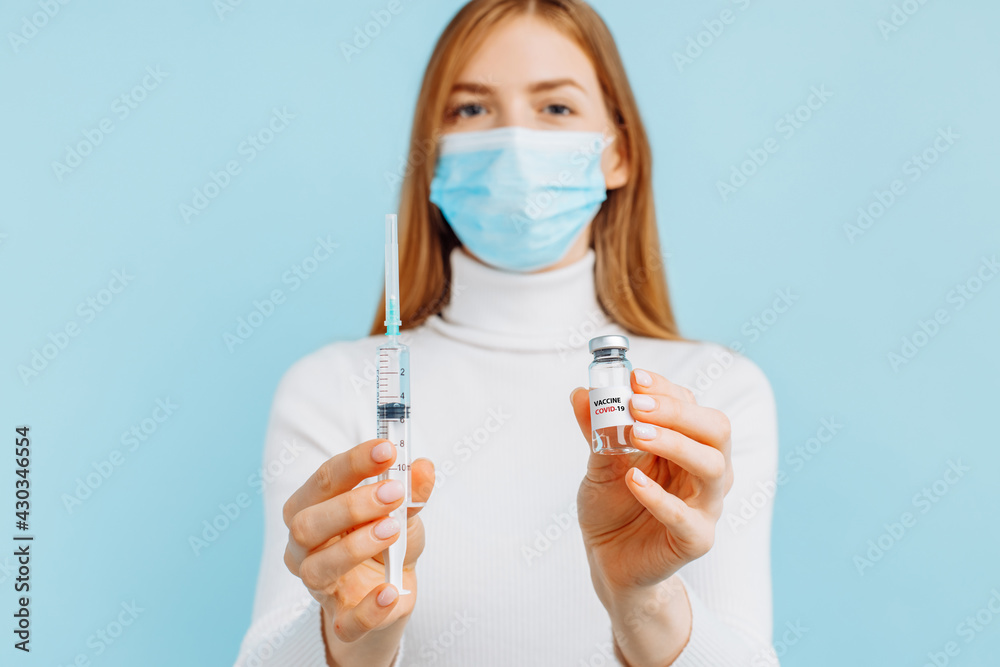 Young woman in a medical mask holding a coronavirus vaccine in a glass bottle and a syringe, on a blue background