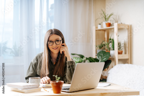 Thinking Freelance woman in glasses with note book typing at laptop and working from home office with plants. Happy girl on workplace at the desk. Distance learning online education and work.