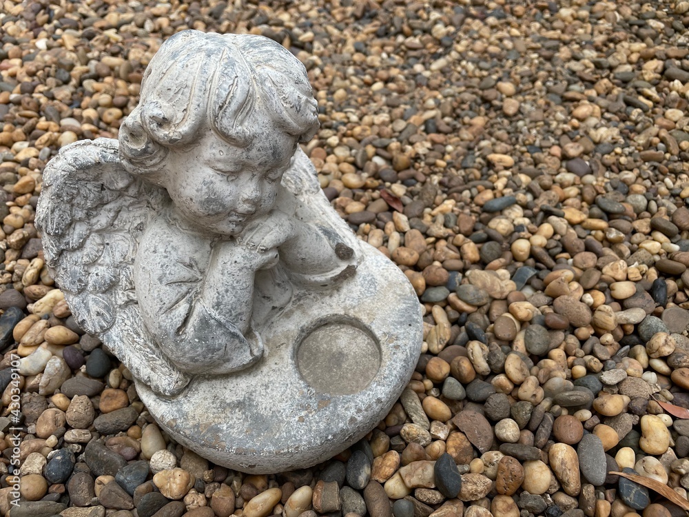 Little cute cupid angel sculpture on river gravel background in the english garden. Little cute cupid angel sculpture on river gravel background in the english garden.