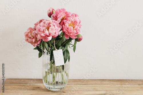Floral still life scene. Pink peonies flowers, bouquet in glass vase on wooden table. Blank gift tag, label mockup. White wall background. Seelective focus. Wedding or birthday celeberation concept.