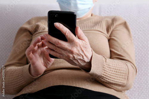 Elderly woman in face mask with smartphone sitting on sofa at home, mobile phone in female hands. Concept of online communication during coronavirus quarantine, sms, social media