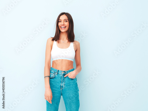 Beautiful smiling woman dressed in white jersey top shirt and jeans. Sexy carefree cheerful model enjoying her morning. Adorable and positive female posing near light blue wall in studio