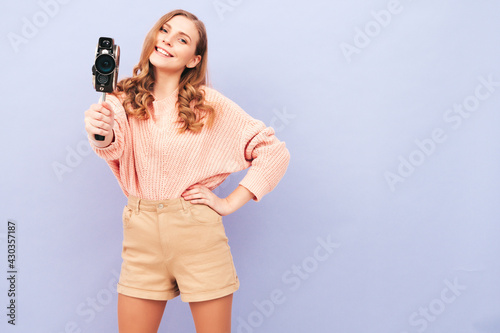 Portrait of young beautiful smiling female in trendy summer hipster clothes. Sexy carefree woman posing near purple wall in studio. Positive model having fun indoors.Holding retro video camera