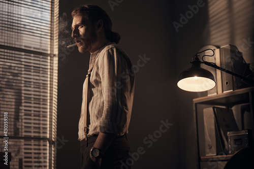 scene of a brutal young detective standing by the window with a thoughtful expression on his face and smoking a cigarette, he is wearing a striped shirt, classic pants with suspenders