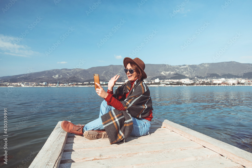 Stylish young beautiful woman traveler sits on the seashore and communicates by video call on a smartphone. She is wearing sunglasses, jeans, a hat. Sunny weather.