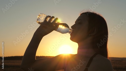 The girl drinks clear cool mineral water from a bottle after training in the park in spring. Young woman drinks water after outdoor workout. Sports in the summer park.