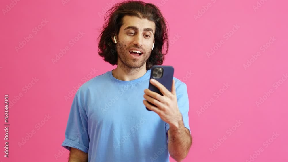 The emotional man in headphones talking by video call on the phone and actively gesturing while standing in a pink studio