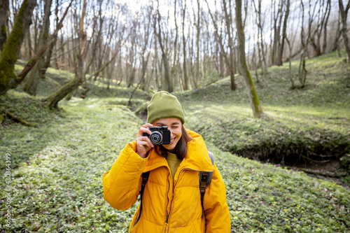 Woman in hiking clothes with backpack covered her face with a vintage camera directed at the camera on green spring forest background. Enjoys of purity and freshness of nature. Travel alone.