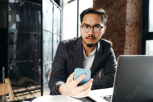 Serious asian businessman sitting at table with laptop and looking at phone while holding it in hand