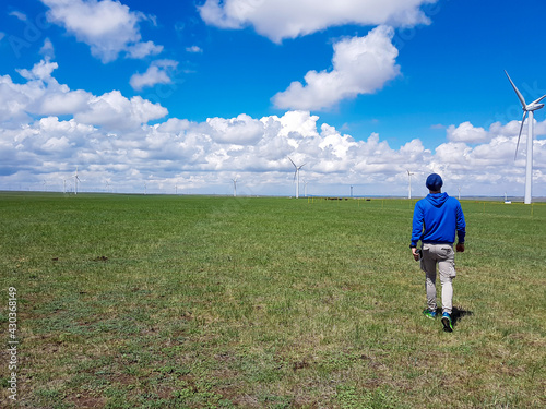 A man walking towards wind turbines build on a vast pasture in Xilinhot in Inner Mongolia. Natural resources energy. Clean energy. Endless grassland. Blue sky with white, thick clouds. Endless horizon
