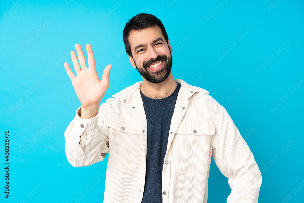 Young handsome man with white corduroy jacket over isolated blue background saluting with hand with happy expression Young handsome man with white corduroy jacket over isolated blue background saluting with hand with happy expression