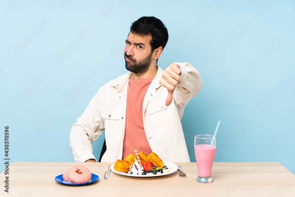 Man at a table having breakfast waffles and a milkshake showing thumb down with negative expression Man at a table having breakfast waffles and a milkshake showing thumb down with negative expression