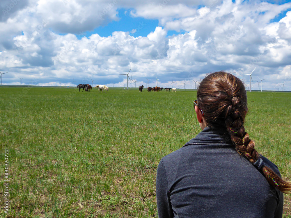 A woman with braided hair watching a heard of horses grazing under wind turbines build on a vast pasture in Xilinhot, Inner Mongolia. Natural resources. Endless grassland. Blue sky with white clouds