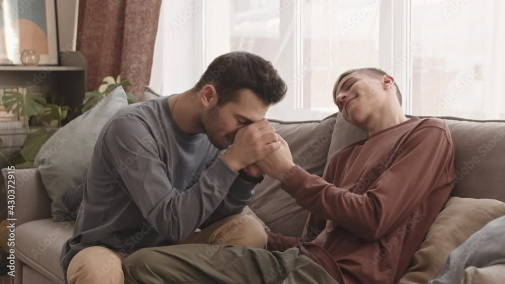 Lockdown of young Caucasian man with dark hair wearing casual clothes sitting on couch and holding hands of his boyfriend sitting nearby Lockdown of young Caucasian man with dark hair wearing casual clothes sitting on couch and holding hands of his boyfriend sitting nearby