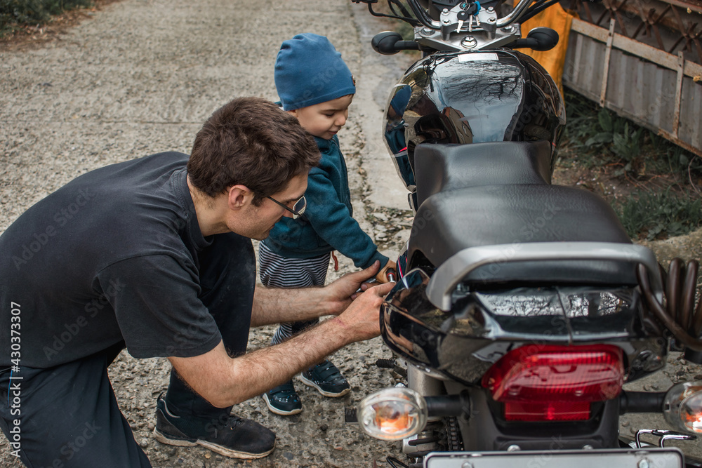 A father and son repair a motorcycle on the street during the day. Communication between father and son.