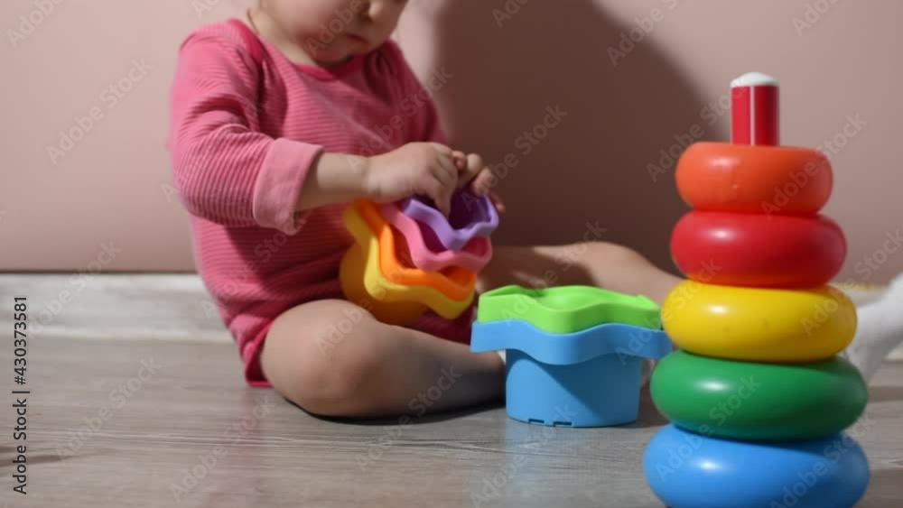Baby girl is playing with stacking cups on the floor and next to it is a stacking rings. She is tryштп to nest all the cups in the right order. Close-up. Baby girl is playing with stacking cups on the floor and next to it is a stacking rings. She is tryштп to nest all the cups in the right order. Close-up.