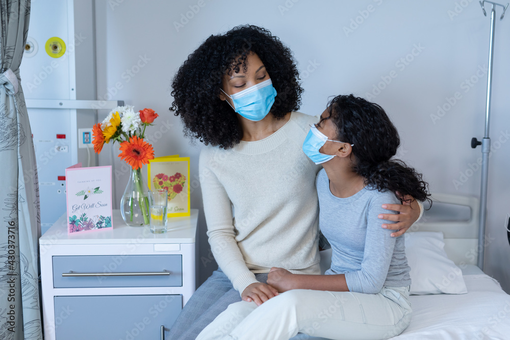 Mixed race mother embracing ill daughter sitting on hospital bed, both wearing face masks Mixed race mother embracing ill daughter sitting on hospital bed, both wearing face masks