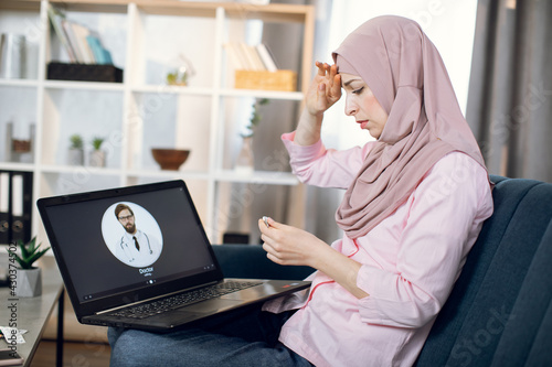 Stay at home concept. Covid prevention. Ill Muslim woman sitting on sofa at home, feeling bad and having fever, holding thermometer and calling to doctor using laptop. Online consultation with doctor