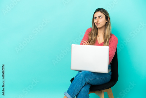 Young caucasian woman sitting on a chair with her pc isolated on blue background making doubts gesture while lifting the shoulders © luismolinero