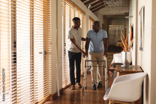 African american young man helping his father to walk with walking frame at home photo