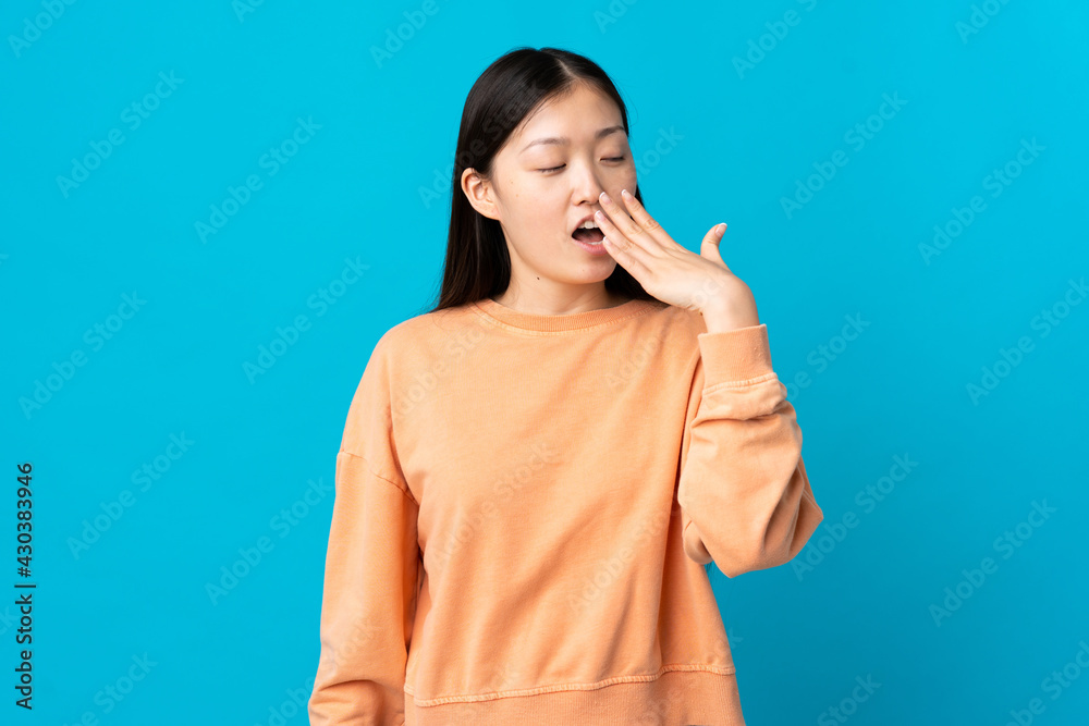 Young Chinese girl over isolated blue background yawning and covering wide open mouth with hand Young Chinese girl over isolated blue background yawning and covering wide open mouth with hand