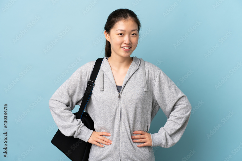 Young sport Chinese woman with sport bag over isolated blue background posing with arms at hip and smiling Young sport Chinese woman with sport bag over isolated blue background posing with arms at hip and smiling