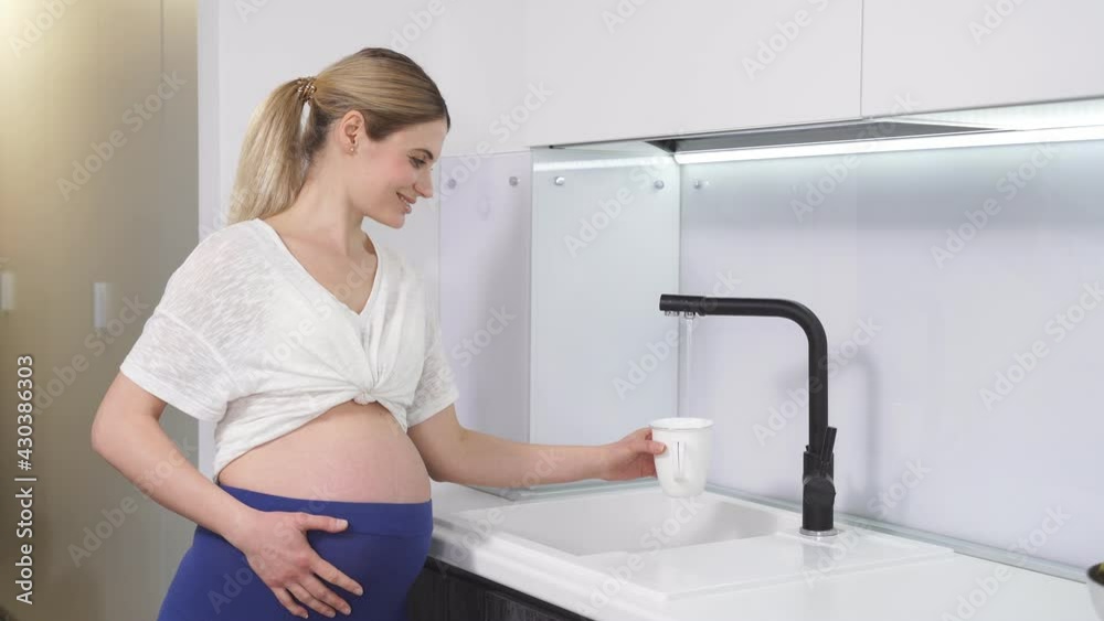 side view of a pregnant woman standing at the tap and pouring water into a drinking glass after a workout side view of a pregnant woman standing at the tap and pouring water into a drinking glass after a workout