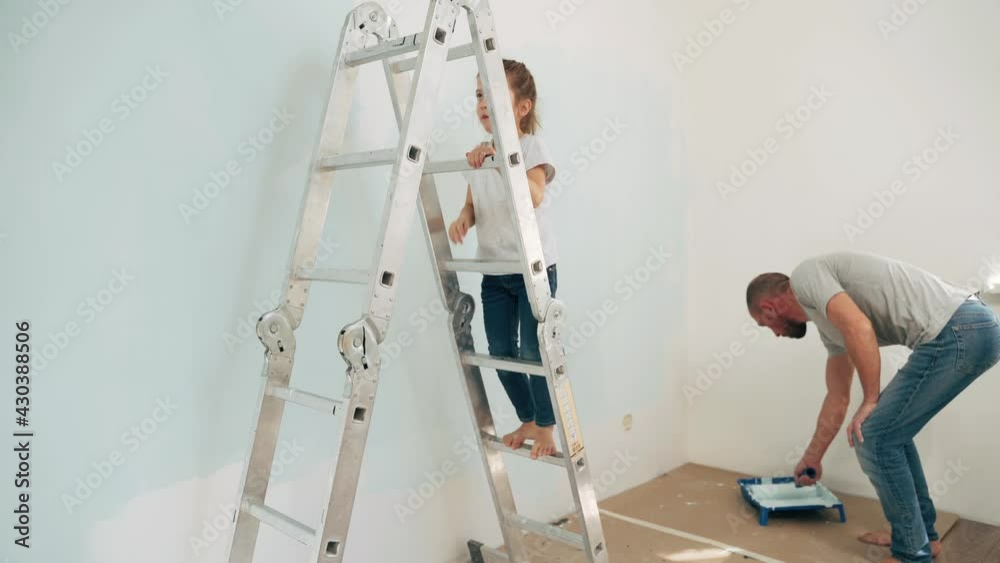 A father and daughter paint a wall A Caucasian man and a girl make repairs A girl standing on a stepladder paints a wall with a roller under the supervision of her father A happy family Side view A father and daughter paint a wall A Caucasian man and a girl make repairs A girl standing on a stepladder paints a wall with a roller under the supervision of her father A happy family Side view