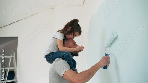 Caucasian man and girl paint the wall Daughter sits on the shoulders of her father and paints the wall with a roller Happy family makes repairs in the room Side view