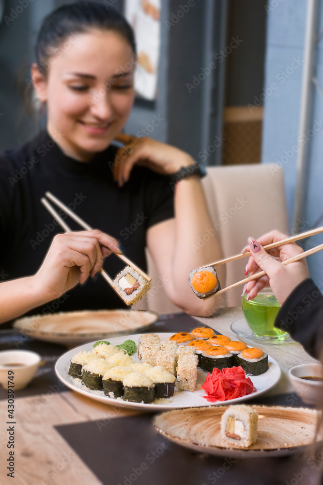 Two women sit at the table and hold sushi with chopsticks over a plate of sushi