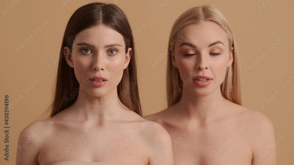 Surprised young half-naked women are looking to the camera standing isolated over a beige background in the studio