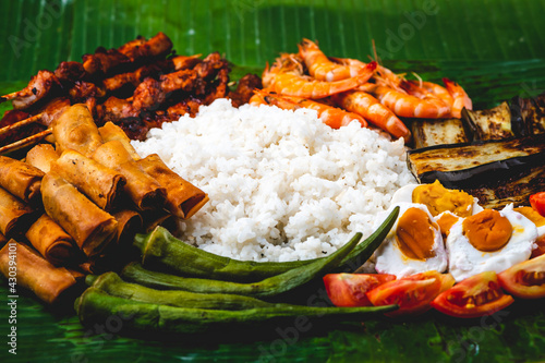 Boodle Fight- White Rice, Lumpiang Shanghai, Salted Egg, Tomato, Boiled Okra, Fried Eggplant, Shrimps, and Pork barbecue (flat lay close up) photo