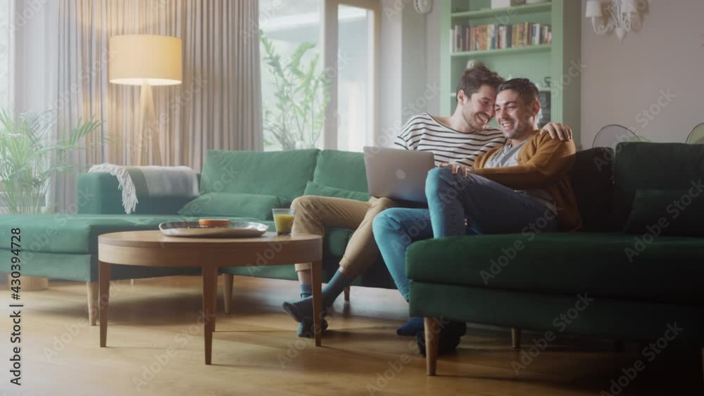 Portrait of Gentle Gay Couple Using Laptop Computer, while Sitting on a Couch in Cozy Stylish Apartment. Adult Boyfriends Online Shopping on Internet, Watching Funny Videos on Streaming Service. Portrait of Gentle Gay Couple Using Laptop Computer, while Sitting on a Couch in Cozy Stylish Apartment. Adult Boyfriends Online Shopping on Internet, Watching Funny Videos on Streaming Service.