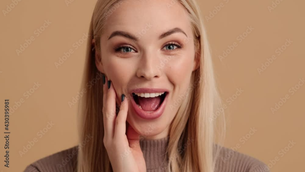 A close-up view of an emotional young blond woman is wondering to the camera standing isolated over a beige background in the studio A close-up view of an emotional young blond woman is wondering to the camera standing isolated over a beige background in the studio