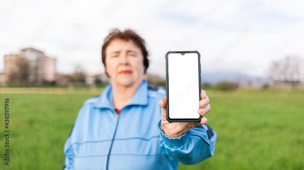 Portrait of an elderly woman in a tracksuit, holding a smartphone with a white screen in her hand. Mock up. Outdoor. The concept of modern technologies for the elderly
