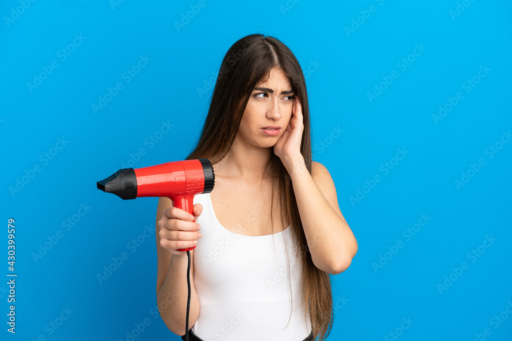 Young caucasian woman holding a hairdryer isolated on blue background with headache Young caucasian woman holding a hairdryer isolated on blue background with headache