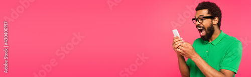 excited african american man in green t-shirt using smartphone isolated on pink, banner