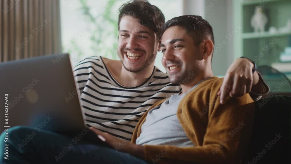 Portrait of Gentle Gay Couple Using Laptop Computer, while Sitting on a Couch in Cozy Stylish Apartment. Adult Boyfriends Online Shopping on Internet, Watching Funny Videos on Streaming Service. Portrait of Gentle Gay Couple Using Laptop Computer, while Sitting on a Couch in Cozy Stylish Apartment. Adult Boyfriends Online Shopping on Internet, Watching Funny Videos on Streaming Service.