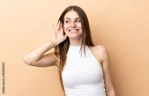 Young caucasian woman isolated on beige background listening to something by putting hand on the ear