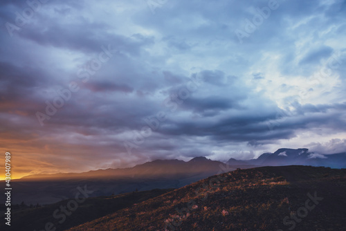 Atmospheric landscape with silhouettes of mountains with pink flowers on hill on background of dawn sky with long orange sun ray on mountain. Vivid scenery with sunset or sunrise of illuminating color