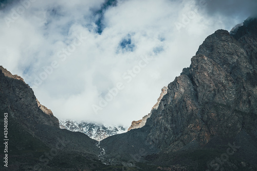 Atmospheric landscape with mountain stream in narrow valley among high rocky mountains. Low clouds on mountain with snow. Beautiful mountain valley with mountain creek. Awesome glen among sharp rocks.