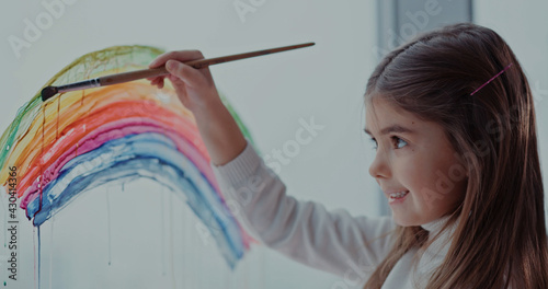 Lovely inspired girl kid drawing colorful watercolor rainbow art on window glass enjoying hobby activity. Cityscape on background.