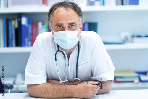 Medico maschio seduto nella postazione del proprio ambulatorio con camice e mascherina chirurgica, guarda con espressione seria