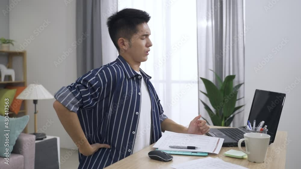 tired asian male doing paperwork is putting down the pen to massage aching lower back and exercising stiff neck with a painful look in the living room at home. tired asian male doing paperwork is putting down the pen to massage aching lower back and exercising stiff neck with a painful look in the living room at home.