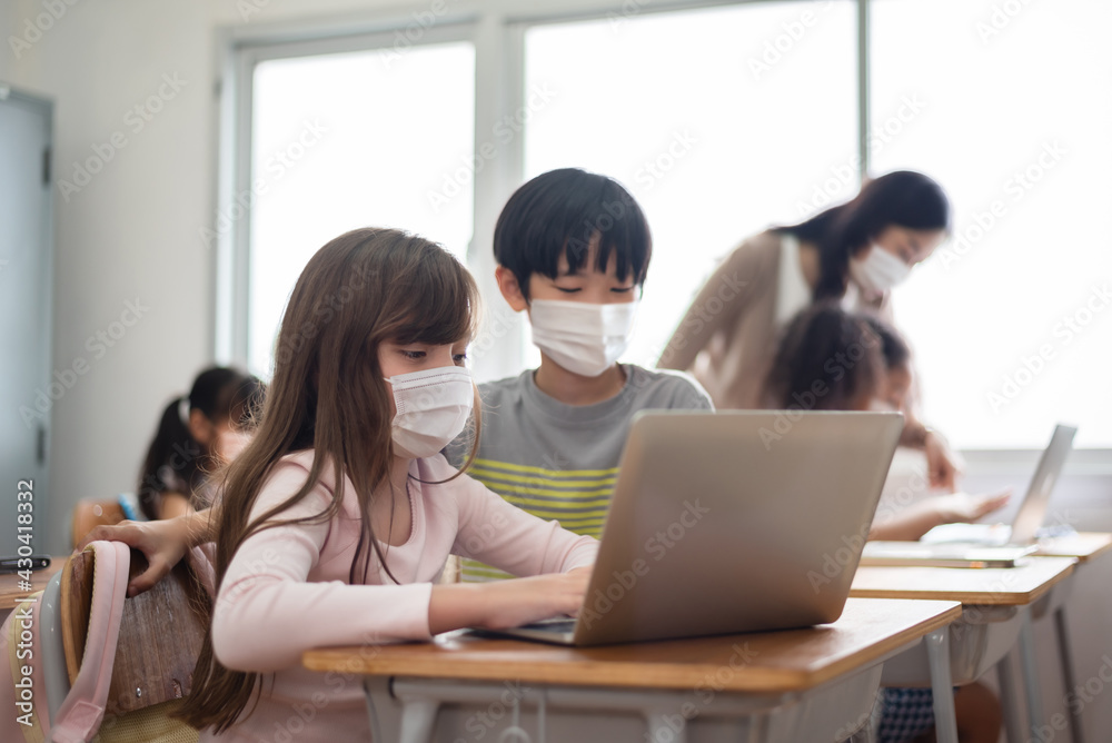 Diverse Multi Ethnic little girls and boys wearing protective face masks using laptop for studying in classroom at primary school Diverse Multi Ethnic little girls and boys wearing protective face masks using laptop for studying in classroom at primary school