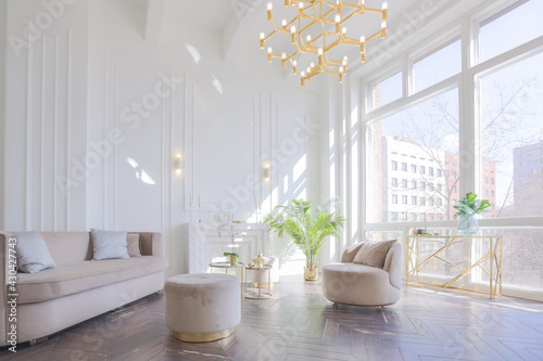 very light and bright interior of luxurious cozy living room with chic soft beige furniture with gold metallic elements, huge window to the floor and wooden parquet