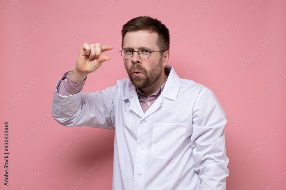 Mocking doctor in round glasses shows small size with fingers and looks into the camera. Pink background. Mocking doctor in round glasses shows small size with fingers and looks into the camera. Pink background.