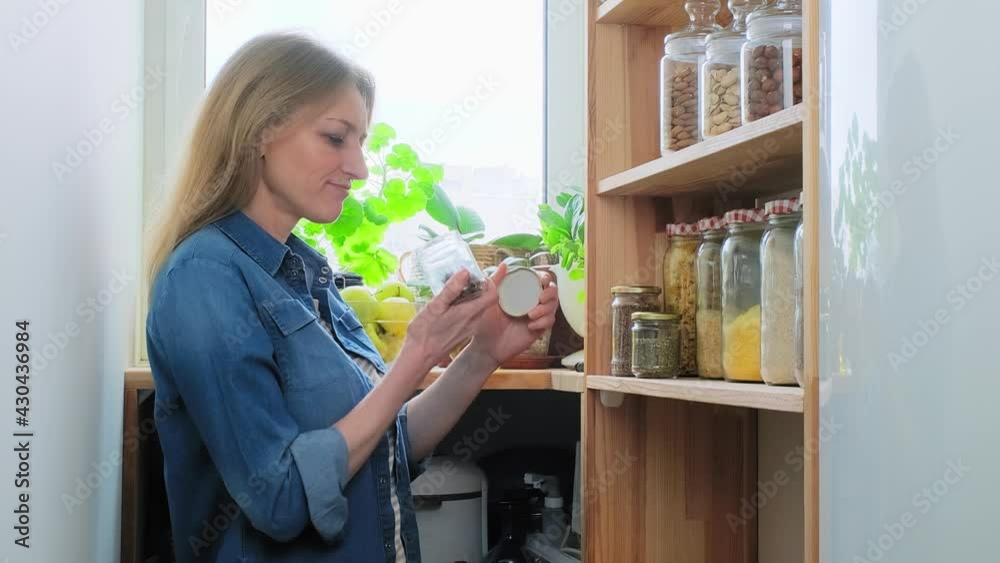 Storing food in pantry, middle-aged woman in kitchen near wooden shelf with jars of cereals Storing food in pantry, middle-aged woman in kitchen near wooden shelf with jars of cereals