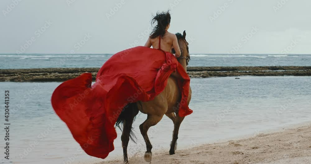 Beautiful woman horseback riding on the beach in flowing red dress, cinematic slow motion
